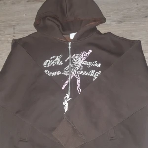 Brun hoodie med tryck och dragkedja - Snygg brun hoodie med huva och dragkedja framtill. Framsidan har ett vitt och rosa tryck med texten 'An Escape from Reality' och en grafisk illustration. Baksidan har ett stort rosa och vitt motiv med döskalle och texten 'The Past is Past'. Perfekt för dig som gillar edgy streetwear.