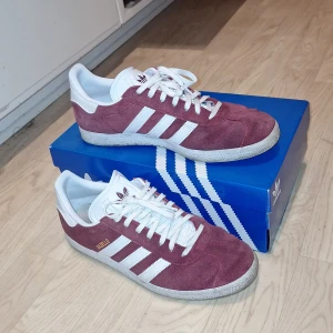 Adidas Gazelle vinröda sneakers - Säljer ett par Adidas Gazelle sneakers i vinrött mocka med vita detaljer och klassiska tre ränder på sidan. Skorna har rund tå, platt sula och snörning. Adidas-logga på plösen och hälpartiet, samt guldfärgad Gazelle-text på sidan. Perfekt för dig som gillar retrostil. Hör av er vid frågor eller funderingar och skicka gärna prisförslag🙌🏼