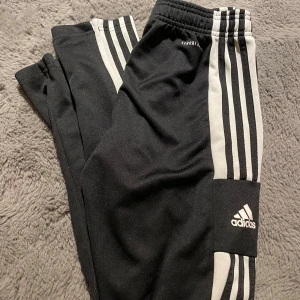 Svarta Adidas mjukisbyxor med vita ränder - Svarta mjukisbyxor från Adidas med klassiska vita ränder längs sidorna och Adidas-logga på benet. Byxorna har resår i midjan och är gjorda i ett mjukt syntetmaterial, perfekt för chill eller träning. Snygg och sportig look med smal passform.