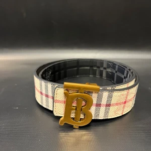 Beige/svart bälte med rutigt mönster - Säljer ett bälte i skinn med klassiskt rutigt mönster i beige, svart och rött på ena sidan och svart rutigt på andra. Bältet har en stor guldfärgad metallspänne med en ikonisk B-logga. Perfekt accessoar för att lyfta din outfit.