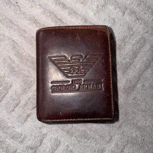 Vintage Brun plånbok från Giorgio Armani - Snygg brun plånbok i äkta skinn från Giorgio Armani med präglad logga på framsidan. Plånboken har flera kortfack, myntfack med knapp och fönster för ID-kort. Klassisk och stilren design som passar perfekt i fickan.