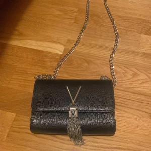 Svart axelväska från Valentino - Snygg svart axelväska från valentino med silverfärgad kedja och ikonisk valentino-logga framtill. Väskan har struktur i materialet, dekorativt hänge med kedjor och praktiskt innerfack med dragkedja. Perfekt för dig som vill ha en stilren och lyxig look.