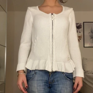 Vit kofta från Odd Molly med volang - Supersöt vit stickad kofta från Odd Molly med volangkant nedtill och snörning vid halsen. Koftan har långärmad passform, ribbade muddar och dragkedja framtill. Perfekt att slänga över en topp för en chill look.