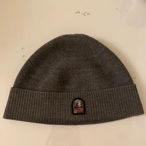 Grå ribbstickad mössa Parajumpers - Snygg grå mössa från Parajumpers med ribbstickad uppvikt kant och klassisk PJS-logga framtill. Mjuk och varm, perfekt för kyliga dagar. Materialet är stickad syntet och designen är enkel men stilren, passar till de flesta jackor och stilar. Bara att skriva vid frågor och priset går såklart att diskutera!