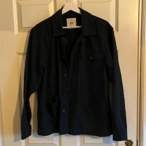 Svart overshirt från Dobber - Stilren svart overshirt från Dobber i storlek S. Jackan har klassisk krage, knäppning framtill med knappar och en bröstficka med lock och knapp. Perfekt lager-på-lager-plagg med enkel och clean look. Jackan är i nyskick aldrig använd, Ny pris 800 mitt pris 450 