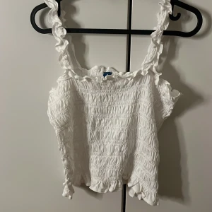 Vit smockad linnetopp från Old Navy - Säljer en vit ribbad linnetopp från Old Navy i storlek M. Toppen har smockad struktur, volangdetaljer vid axelbanden och längs kanten, samt ärmlös design. Perfekt för sommarens alla tillfällen och superfin till både jeans och kjol. 