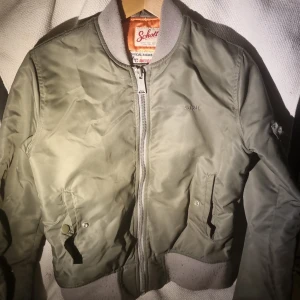 Olivgrön bomberjacka från Schott - Säljer en olivgrön bomberjacka från Schott med orange satin-foder och grå ribbade muddar. Jackan har dragkedja framtill, fickor med tryckknapp och en ärmficka med patch. Klassisk college-vibe och riktigt snygg passform.