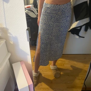 Blå blommig långkjol med slits - Supersnygg blå långkjol med vitt blommönster och hög slits på sidan. Kjolen har en smickrande passform och är perfekt för dig som gillar en feminin och trendig stil. Lätt och flowy material som känns skönt mot huden.