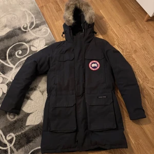 Canada Goose jacka - Säljer en svart parkas från Canada Goose med ikonisk logga på bröstet och stor huva med fuskpäls.                                                                 * Priset kan diskuteras vid snabba affärer