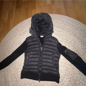 Svart pufferjacka från Moncler - Äkta.Säljer en svart pufferjacka från Moncler med stickade ärmar och huva. Jackan har dragkedja framtill, två fickor med knappar och en extra ficka med dragkedja på ärmen. Perfekt för dig som vill ha en snygg och sportig look.