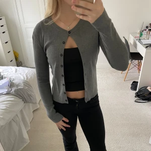 Grå kofta från H&M med knappar - Säljer en stilren grå kofta från H&M i storlek S. Koftan har rund halsringning, långa ärmar och svarta knappar hela vägen framtill. Materialet är mjukt och stickat, perfekt att slänga över en topp. Enkel och clean look som funkar året runt.