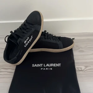 Svarta Saint Laurent sneakers - Svarta sneakers från Saint Laurent med vit logotyp på sidan och klassisk rund tå. Skorna har snörning och en beige platt sula. Materialet är skinn och textil, och de kommer med original dustbagoch box. Perfekt för dig som gillar stilrena designer.