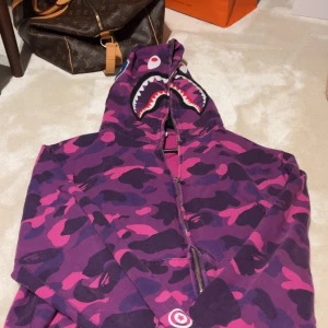 Lila camo hoodie från Bape Shark - Säljer en Riktig Bape Shark hoodie i lila och rosa camomönster. Tröjan har dragkedja hela vägen, huva med hajansikte och broderade detaljer. köpt i USA för 5000 kr, använt runt 4 gånger, säljer pga: tyvärr inte min stil 💗