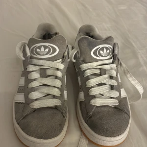 Gråa Adidas sneakers med vit logga - Gråa Adidas sneakers i mocka med klassiska vita ränder på sidorna och vit Adidas logga på plösen. Skorna har vita snören och en vit sula med gummidetaljer. Snygg och enkel design som passar till det mesta.