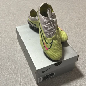 Nike Phantom GX Elite fotbollsskor gul - Nike Phantom GX Elite fotbollsskor i neon-gult och vitt med svarta och rosa detaljer. Skorna har snörning, platt sula med dobbar och är tillverkade i syntetmaterial. Swoosh-logga i svart och rosa på sidan. Levereras med matchande gympåse och original skokartong. Säljes p.g.a att jag slutade med fotbollen och hann knappt använda skorna.