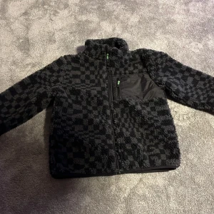 Svart jacka - Mysig svart teddyjacka från H&M med rutigt mönster i olika nyanser av svart och grått. Jackan har hög krage, dragkedja framtill och en bröstficka i nylon med grön detalj på dragkedjan. Perfekt för kyliga dagar och riktigt skön att bära. Kan diskutera pris