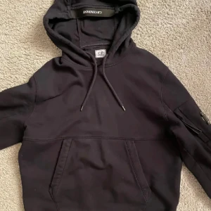 Svart hoodie från C.P. Company - Svart hoodie från C.P. Company med klassisk linsdetalj på ärmen, snörning i huvan och stor magficka. Tröjan har lång ärm och normal passform, tillverkad i mjuk bomull. Perfekt för dig som gillar streetwear och snygga detaljer. Går ner i pris snabb affär och tar byten