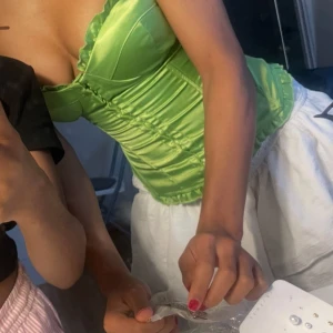 Grön korsett från Charlotte Russe - Snygg grön korsett från Charlotte Russe i glansigt material. Den har snörning bak och volangdetaljer upptill och nedtill. Perfekt för att ge en elegant touch till din outfit.