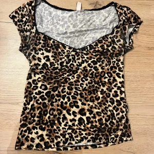 Leopardmönstrad topp från Nelly - Säljer en kortärmad topp från Nelly i storlek S med leopardmönster i brunt, svart och beige. Toppen har v-ringad hals med svart spetskant och är tillverkad i mjukt syntetmaterial. Perfekt för dig som gillar djurmönster och vill sticka ut.