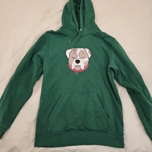 Grön hoodie från loop snus  - Säljer en grön hoodie med en stor ficka framtill och snörning i huvan. På bröstet finns ett tryck av en bulldog med röd taggkrage. Perfekt för dig som gillar streetstyle och vill ha något bekvämt och snyggt.