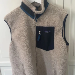 Beige fleeceväst från Patagonia - Säljer en beige fleeceväst från Patagonia med marinblå detaljer och hög krage. Västen har en bröstficka med dragkedja och klassisk Patagonia-logga. Perfekt för lager-på-lager och outdoor-vibe. Stängs med dragkedja framtill och har mjukt, teddy-liknande material.