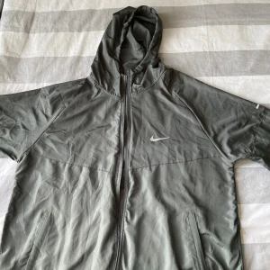 Grå Nike vindjacka med huva - Nike Running vindjacka i grått med huva och hel dragkedja framtill. Jackan har två fickor med dragkedja och en stilren Nike-logga på bröstet. Lätt och smidig modell, perfekt för löpning eller utomhusaktiviteter.
