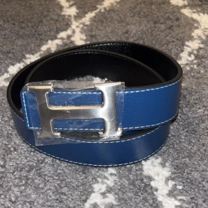 H-bälte  - Snyggt bälte i blått och svart skinn med ikoniskt,H-spänne i silverfärgad metall. Bältet har stilrena, kontrastsömmar och klassisk design som lyfter, vilken outfit som helst. Strl 110cm