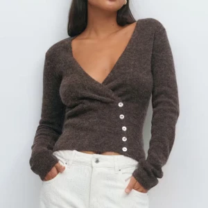 Brun omlottkofta med v-ringning - Snygg brun omlottkofta med v-ringad hals och sex dekorativa knappar framtill. Koftan är stickad och har långärmad passform som sitter tajt mot kroppen. Perfekt för dig som gillar en stilren och trendig look med lite edge.