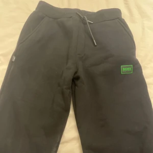 Svarta mjukisbyxor från BOSS - Svarta mjukisbyxor från BOSS med grön logga på benet och snörning i midjan. Byxorna har ribbade muddar och är tillverkade i mjukt bomullsmaterial. Perfekta för en chill och avslappnad stil.