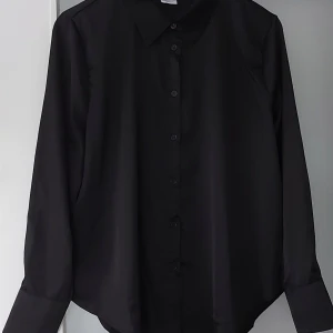Svart silk skjorta från Åhléns - Stilren svart skjorta från Åhléns i storlek L. Aldrig använt. Skjortan har klassisk krage, knappar hela vägen och långa ärmar med knapp vid manschetten. Materialet är väldigt skönt och det har en lätt glansig finish som ger en snygg look. 