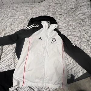 Manchester United träning/regn jacka  - Snygg Manchester United träning/regn jacka från Adidas med svartvita ärmar. Jackan har huva, dragkedja och klubbmärke på bröstet. Perfekt för dig som vill ha sportig stil och visa vilket lag du håller på.