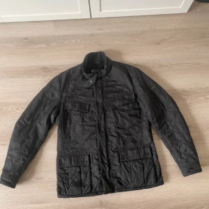 Svart quiltad jacka från Barbour - Svart quiltad jacka från Barbour International med klassisk krage, tryckknappar och dragkedja. Mycket bra skick storlek L.