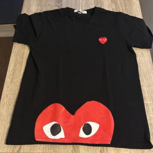 Svart Comme des Garçons Play t-shirt - Svart t-shirt från Comme des Garçons Play med ikoniskt rött hjärta med ögon broderat på bröstet och stort tryck av samma hjärta längst ner. Klassisk rund hals och korta ärmar. Materialet är mjuk bomull som känns skön mot huden.