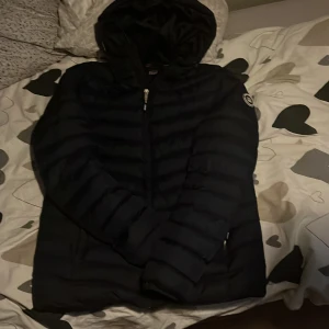 Mörkblå pufferjacka från Vinson - Säljer en mörkblå pufferjacka från Vinson med huva och snyggt märke på ärmen. Jackan har horisontella quiltade partier, dragkedja framtill och två sidofickor. Perfekt för dig som vill ha en stilren och varm jacka med modern look.