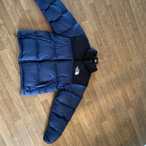 Blå dunjacka från The North Face - Säljer en blå och svart dunjacka från The North Face med logga på bröstet och ryggen. Jackan har hög krage, dragkedja framtill och två sidofickor. Perfekt för kalla dagar och riktigt snygg streetstil.