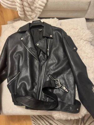 Svart skinnjacka från Missguided - Säljer en svart skinnjacka från Missguided i klassisk bikerstil. Jackan har silvriga dragkedjor, bred krage, axeldekorationer och ett bälte i midjan. Snygg och edgy look som passar perfekt till jeans eller klänning.
