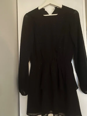 Svart glittrig klänning! - Säljer en svart kortklänning med diskret glittrigt tyg och långa ärmar. Klänningen har en markerad midja och lätt utställd kjol, vilket ger en snygg siluett. Perfekt för dig som vill ha en enkel men ändå festlig look. Från gina i storlek 158/164, i nyskick💕