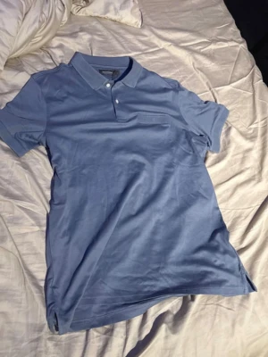 Blå pikétröja från Dressmann - Säljer en stilren blå pikétröja från Dressmann i mjuk bomull. Tröjan har klassisk krage med två knappar och en diskret bröstficka med mesh-detalj. Perfekt till jeans eller shorts för en clean look.