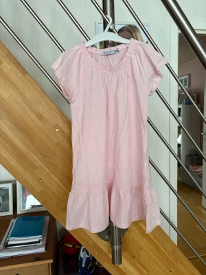Ljusrosa kort klänning från 157 - Supersöt ljusrosa klänning från 157 med kort ärm och volang nertill. Klänningen har en rundad hals med resår och är gjord i mjuk bomull, perfekt för varma dagar. Enkel och stilren design som är lätt att matcha med sneakers eller sandaler.