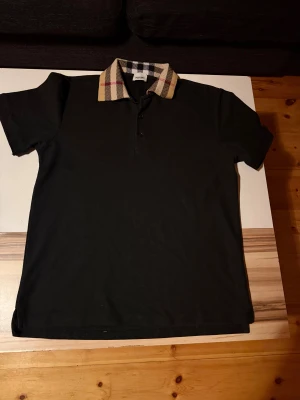  Burberry pike med rutig krage - Snygg svart piké från Burberry med klassisk rutig krage i beige, svart, vitt och rött. Modellen har korta ärmar och knäppning framtill. Perfekt för dig som vill ha en stilren look med en twist från det ikoniska märket.