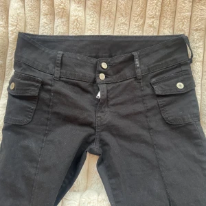 Svarta bootcut byxor med fickor - Säljer ett par svarta bootcut byxor med dubbla knappar i midjan och stora fickor framtill med lock och knapp. Byxorna har en utsvängd passform nertill och är sydda i ett mjukt bomullstyg. Perfekta för dig som gillar retrovibbar och vill ha något som sticker ut. Har ett hål, se sista bilden. Står ingen storlek eller märke men passar xs/s
