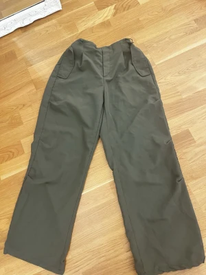 Olivgröna vida byxor med fickor - Säljer ett par olivgröna byxor med bred passform och stora fickor framtill. Byxorna har hög midja och raka ben, perfekta för en avslappnad och trendig look. Materialet känns slitstarkt och passar till många olika stilar.