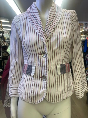 Randig kavaj i bomull med knappar - Snygg vit kavaj med röda och svarta ränder, tillverkad i bomull. Kavajen har två stora knappar framtill, fickdetaljer med flerfärgade ränder och markerad krage med broderade sömmar. Långärmad och figurnära passform, perfekt för en trendig look.