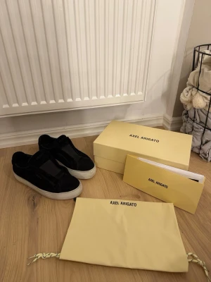 Svarta sneakers från Axel Arigato - Säljer ett par svarta sneakers från Axel Arigato i mocka med vit sula. Skorna har minimalistisk design utan snörning och elastiskt band över vristen för enkel påtagning. Kommer med originalkartong, dustbag och klistermärken.