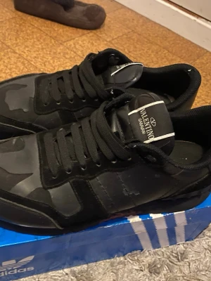 Valentino Garavani svarta sneakers - Säljer ett par svarta Valentino Garavani sneakers med coolt camouflagemönster. Skorna har snörning och är tillverkade i en mix av mocka och skinn, vilket ger en lyxig känsla. Perfekta för dig som vill ha något unikt och stilrent i garderoben.