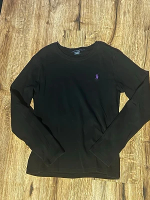 Svart långärmad tröja Ralph Lauren - Svart långärmad tröja från Ralph Lauren Sport i 100% bomull. Klassisk passform med rund hals och lila broderad logga på bröstet. Perfekt till jeans eller kjol för en clean och stilren look. Står L men passar S. Förmodligen pgaav feltvätt