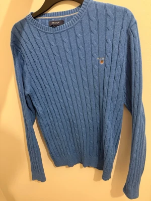 Blå kabelstickad tröja från Gant - Säljer en blå kabelstickad tröja från Gant med rund halsringning och diskret logga broderad på bröstet. Tröjan har långa ärmar och ribbade muddar vid ärmslut och nederkant. Perfekt för dig som gillar klassisk och stilren look.