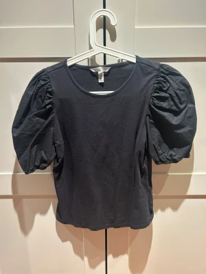 Svart topp med puffärmar - Säljer en svart topp med riktigt snygga puffärmar och rund halsringning. Toppen är i mjuk bomull och har en enkel, stilren design som ändå sticker ut tack vare de voluminösa ärmarna. Perfekt att styla med jeans eller kjol för en trendig look.