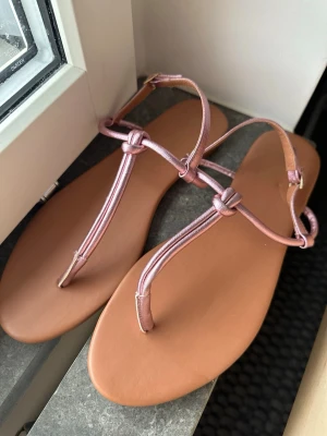 Rosa metallic sandaler från H&M - Säljer ett par helt nya stilrena sandaler från H&M med tunna remmar i metallicrosa och snygga knutdetaljer. Sulan är i en varm beige ton och sandalerna har öppen tå och justerbart spänne vid ankeln. 