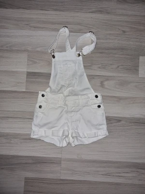 Vita korta hängselshorts H&M - Snygga vita hängselshorts från H&M i mjuk bomull. Klassisk design med justerbara axelband, fickor fram och bak samt knappar i sidorna. Perfekta till sommaren och lätta att matcha med en t-shirt eller linne.